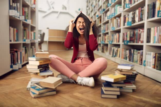 A young woman is sitting at a desk in a library, surrounded by books, with a laptop open in front of her. She is focused on the screen, with a determined expression on her face, symbolizing dedication and the pursuit of knowledge.