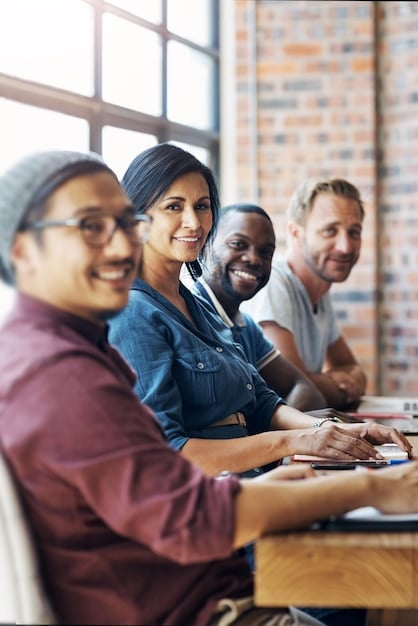 A medium shot of a diverse group of people participating in a workshop. They are gathered around tables, actively engaged in discussions and writing notes. The atmosphere is collaborative and supportive. The workshop space is bright and modern, with white walls and colorful accents. There's a whiteboard in the background with brainstorming ideas written on it.