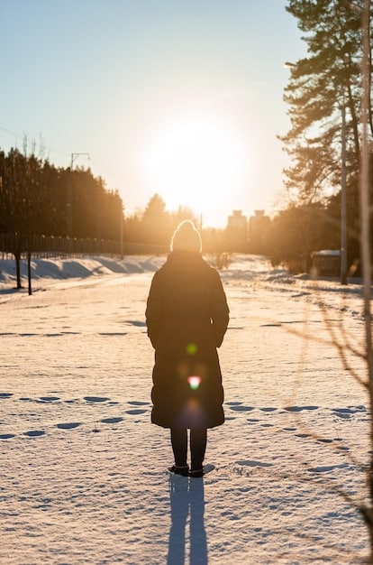 A person bundled up in warm winter clothing, standing outside in a snowy landscape, looking up at the slightly visible sun.