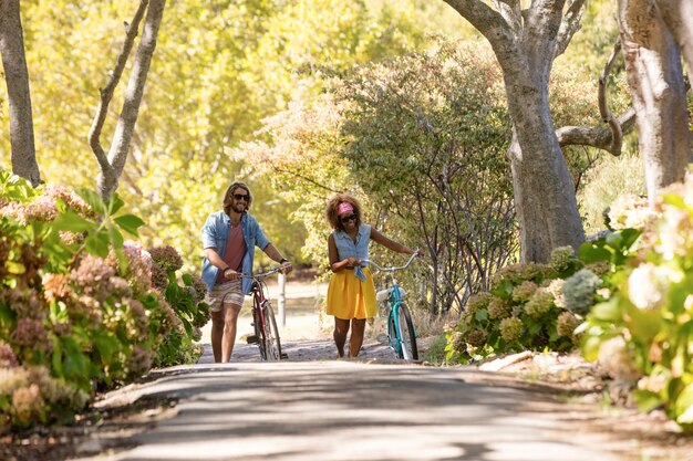 A diverse group of people riding bicycles together on a paved bike path through a park. The trees are lush and green, and the sun is shining.