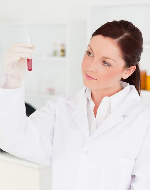 A brightly lit laboratory setting with a female scientist in her 40s analyzing a vial of clear liquid. She is wearing a lab coat and safety glasses, looking attentively at the sample. Scientific equipment and beakers are visible in the background, indicating a focus on research and development.