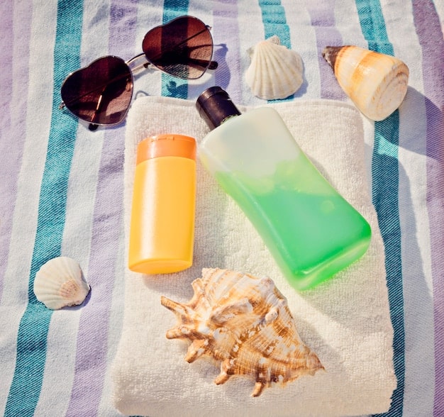 A selection of men's sunscreens (lotion, stick, spray) arranged on a beach towel with a beach background. The scene suggests sun protection as part of an active, outdoor lifestyle.